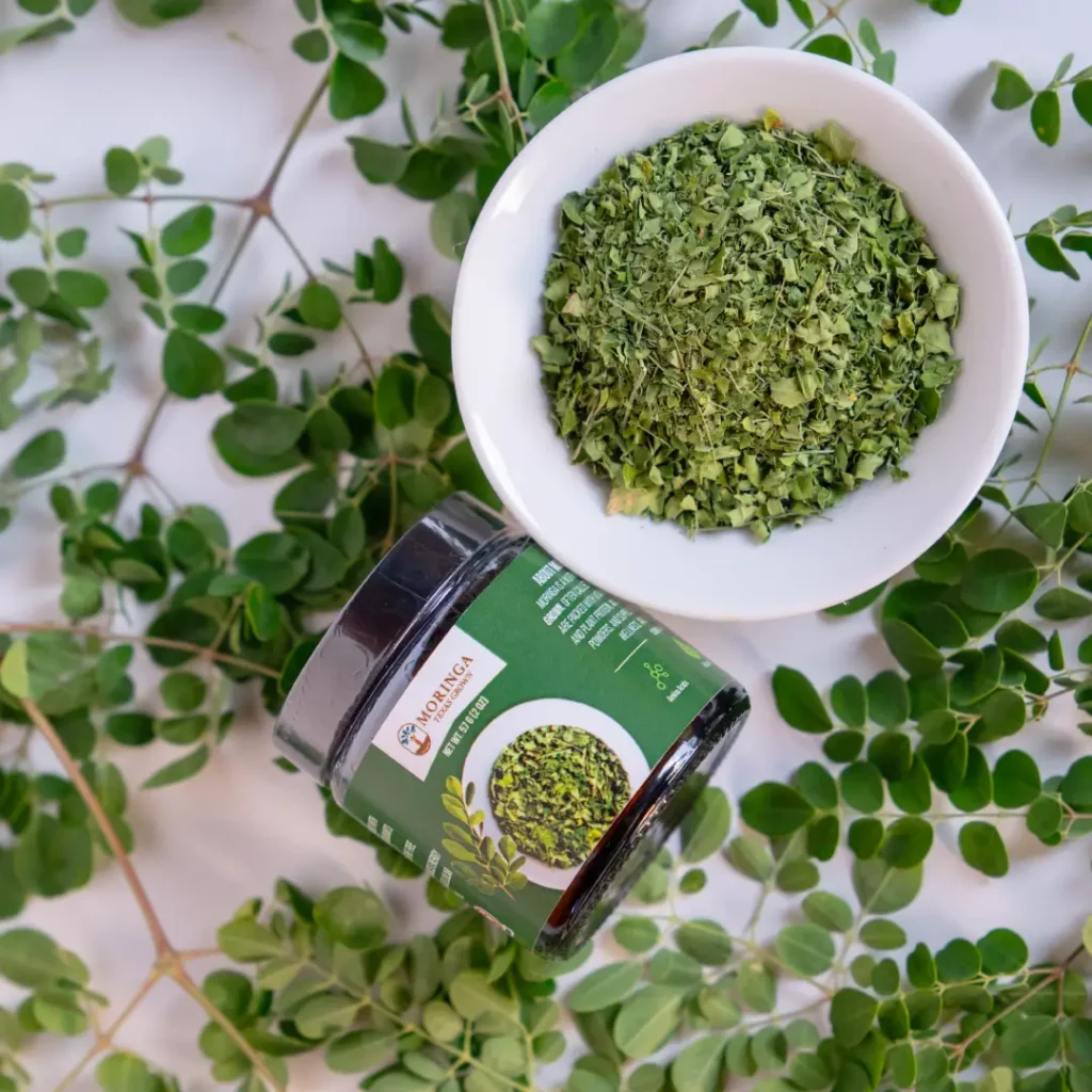 Dried moringa leaves in a white bowl beside a Moringa Texas Grown container surrounded by fresh moringa leaves.